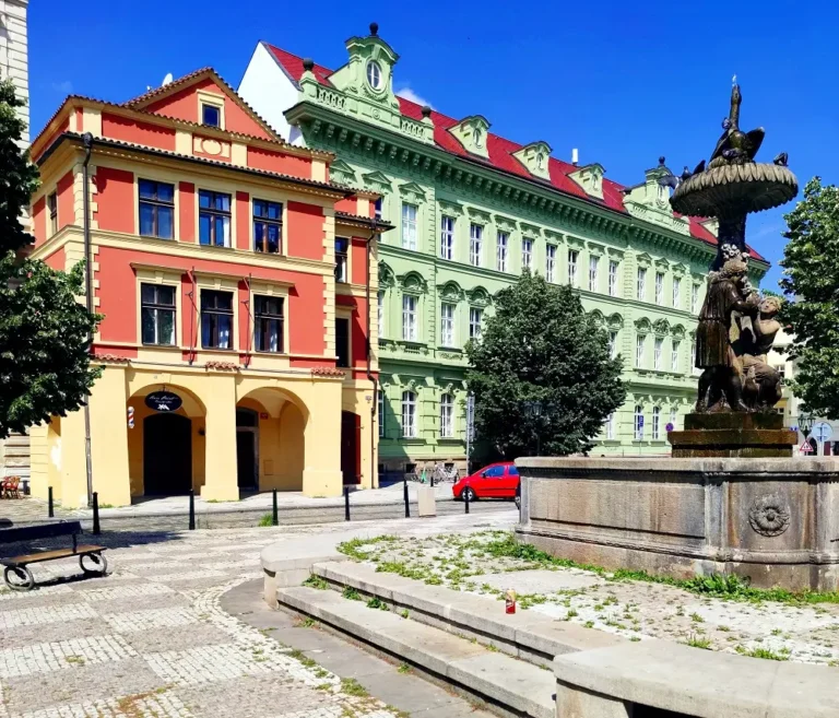 дом U Tří stupňů с красным фасадом в Праге, вид на площадь со старинным фонтаном и скульптурами в солнечный день